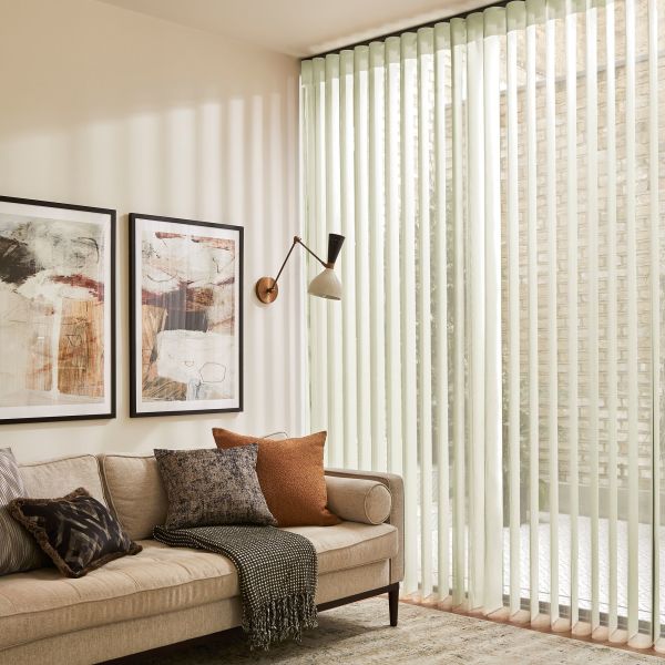 Pale fabric vertical blinds cover a floor‑to‑ceiling sliding glass door, slats angled to filter soft daylight, casting stripes; cozy living room with beige sofa, cushions, wall lamp and framed art.