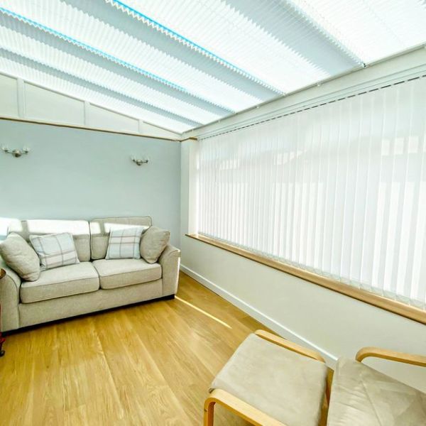 Vertical white blinds cover the side window, while pleated translucent roof blinds cross the ceiling, both closed and softly diffusing daylight; a small conservatory with a pale sofa and wooden floor.