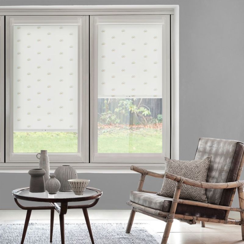White roller blinds with tiny rainbow motifs, fitted inside a two‑pane window — left fully lowered, right partially raised, softly filtering daylight; living room with gray walls, rattan chair and coffee table.