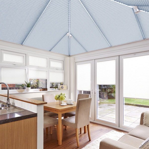Blue pleated roof blinds fitted to the pitched conservatory ceiling, with white pleated window and door blinds lowered, filtering soft daylight over a dining area, kitchen sink, and garden view.