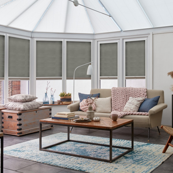Gray pleated cellular blinds, lowered inside white-framed conservatory windows and filtering sunlight; situated above a beige sofa and wooden coffee table in a softly lit, pastel-accented living room.