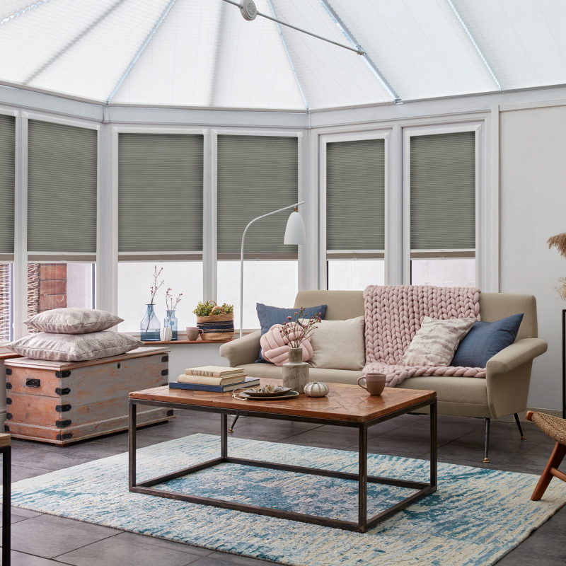 Gray pleated cellular blinds, lowered inside white-framed conservatory windows and filtering sunlight; situated above a beige sofa and wooden coffee table in a softly lit, pastel-accented living room.