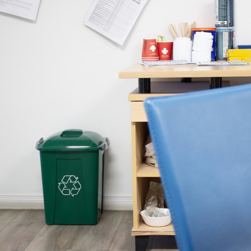 green plastic recycling bin with a white mobius loop icon printed on the side with lockable lid for office recycling sitting on the floor beside a desk