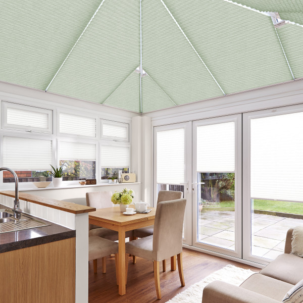 Light-green pleated roof blinds cover the pitched conservatory ceiling, with white pleated blinds on windows and French doors, closed and diffusing daylight over a dining area facing a patio garden.