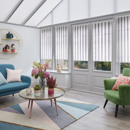 Striped vertical blinds, fully lowered on tall glazed doors and windows, pale-striped; filtering soft daylight in a conservatory with glass roof, teal sofa, green armchair, coffee table and pastel rug.
