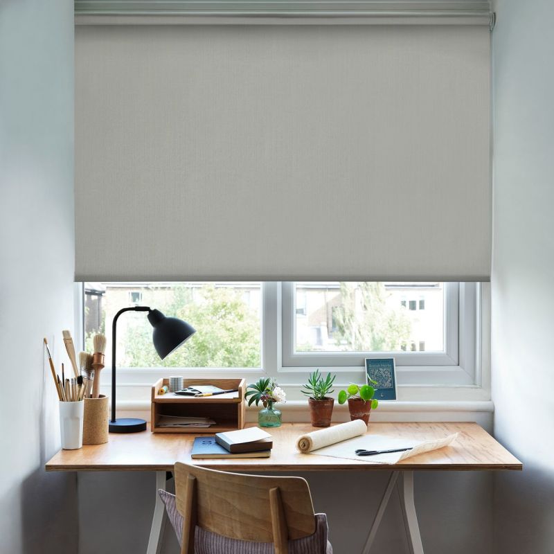 A light-gray roller blind, lowered almost fully and softly diffusing daylight, covers a double window above a tidy wooden desk with lamp, brushes, notebooks and potted plants.