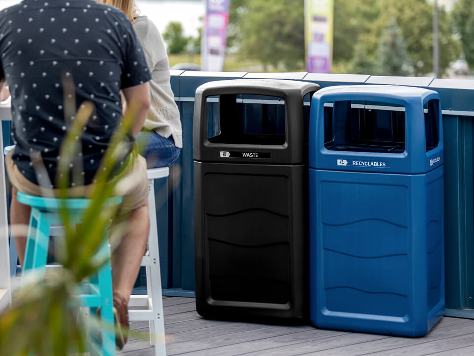 black and blue waste and recyclables bins made from 100% PCR material outdoors at patio bar with people sitting at table