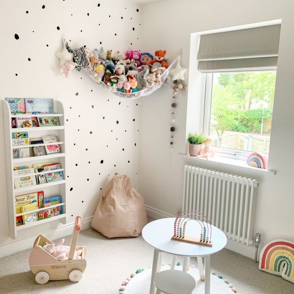 Grey Roman fabric blind (folded tiers) partially lowered, filtering daylight through the window; bright children's nursery with white polka-dot walls, stuffed-animal hammock, bookshelf, beanbag, small table, garden view.
Text found: Maeva
Text found: Dear Zoo