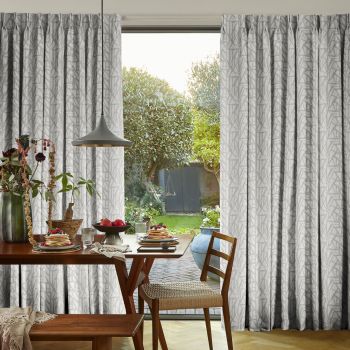 Floor‑length pleated gray patterned curtains, drawn aside, framing a glass door and filtering soft daylight into a dining nook with wooden table, pendant lamp and garden view.