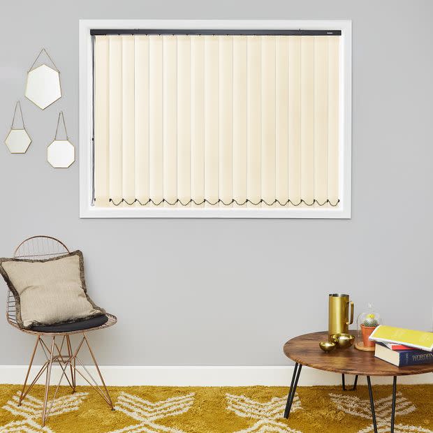 Cream vertical blinds fully closed across a window, slatted with weighted chains; filtering soft daylight into a grey-walled living room with wire chair, cushion, round wooden table and mustard rug.