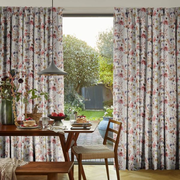 Floral pleated full-length curtains patterned with pink, mauve and grey blossoms frame a sliding glass door, partially drawn to filter light, overlooking a sunlit backyard garden and a breakfast table.