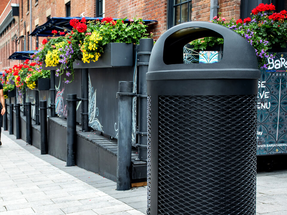 all black waste container with metal body and plastic domed lid perfect for keeping contents dry sitting beside an outdoor downtown restaurant patio