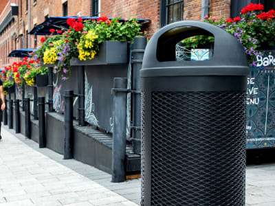 all black waste container with metal body and plastic domed lid perfect for keeping contents dry sitting beside an outdoor downtown restaurant patio