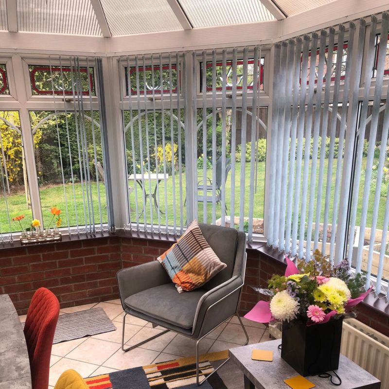 Pale-gray vertical blinds hang across wraparound conservatory windows, slats partially angled and filtering daylight; interior shows a tiled sunroom with an armchair, cushion, small table and garden view.