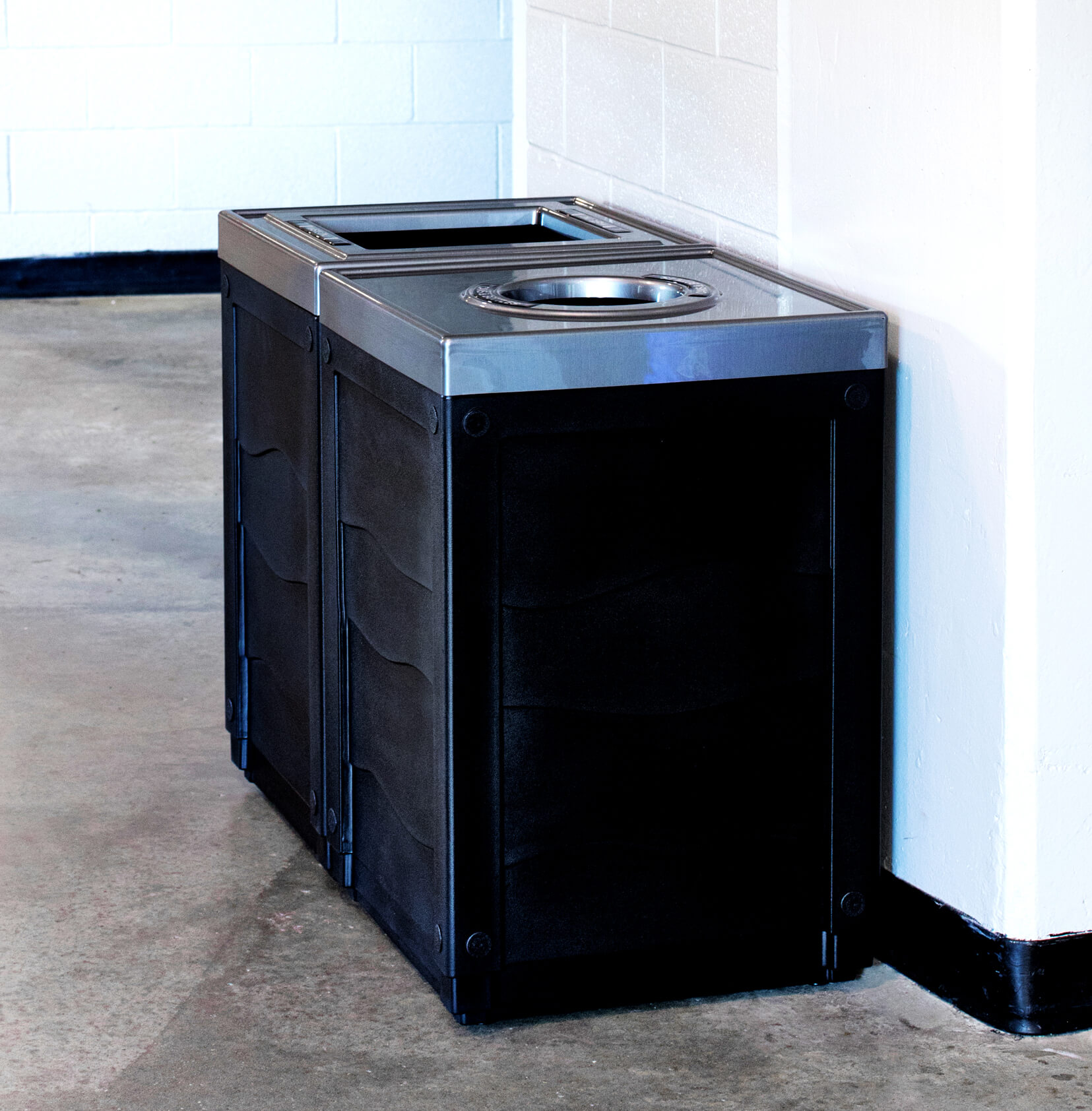 double waste and cans and bottles bins with black bodies against a wall in the hallway of an arena