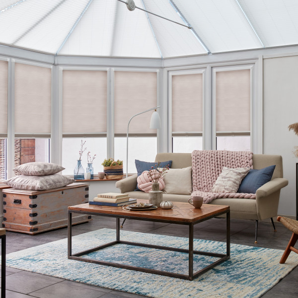 Pleated beige cellular blinds, lowered across bay windows and conservatory panels, closed and diffusing daylight; conservatory living area with neutral sofa, chunky pink throw, wooden coffee table and blue rug.
