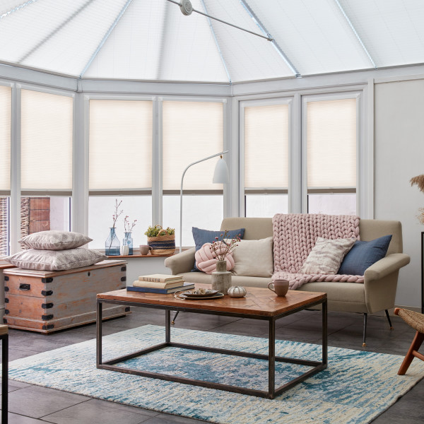 Pleated cellular blinds, fully lowered on bay windows with light‑filtering opaque fabric and narrow horizontal pleats; filtering soft daylight into a conservatory‑style living room with sofa, coffee table, rug.