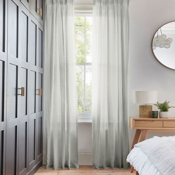 Sheer floor-length pale-gray curtains, pleated and hung at a rod, filtering soft daylight through a tall window; bedroom context with dark paneled wardrobe left, wooden nightstand and round mirror.