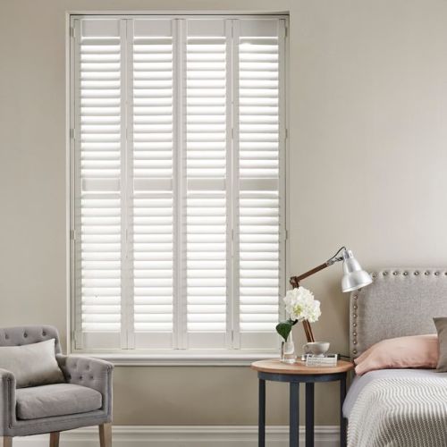White plantation shutters covering a tall window, horizontal slats angled to filter bright daylight, in a softly lit bedroom corner with upholstered bed, side table, lamp and armchair.