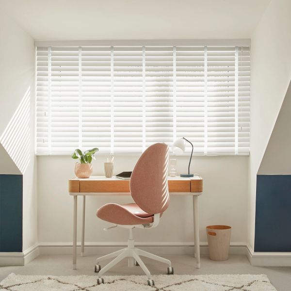 White horizontal blinds across a wide alcove window, closed and filtering bright daylight into striped shadows; small home workspace with wooden desk, pink swivel chair, table lamp, potted plant, walls.
