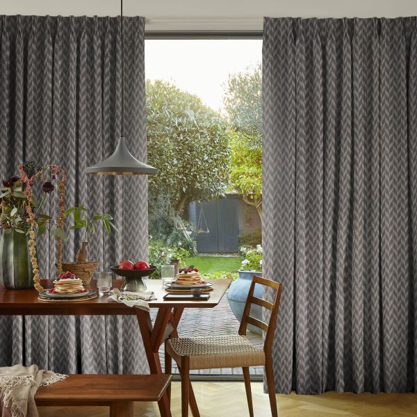 Grey patterned pleated curtains, ceiling-mounted and drawn aside to reveal the patio door, framing a sunlit backyard; a wooden dining table set for breakfast with pancakes, pendant lamp and chairs.