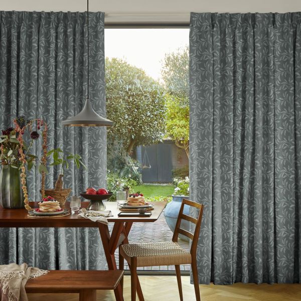Floor-length grey patterned curtains, pleated and partially drawn to reveal a central glass door, filtering soft daylight, in a dining room with wooden table, pendant lamp and leafy garden beyond.