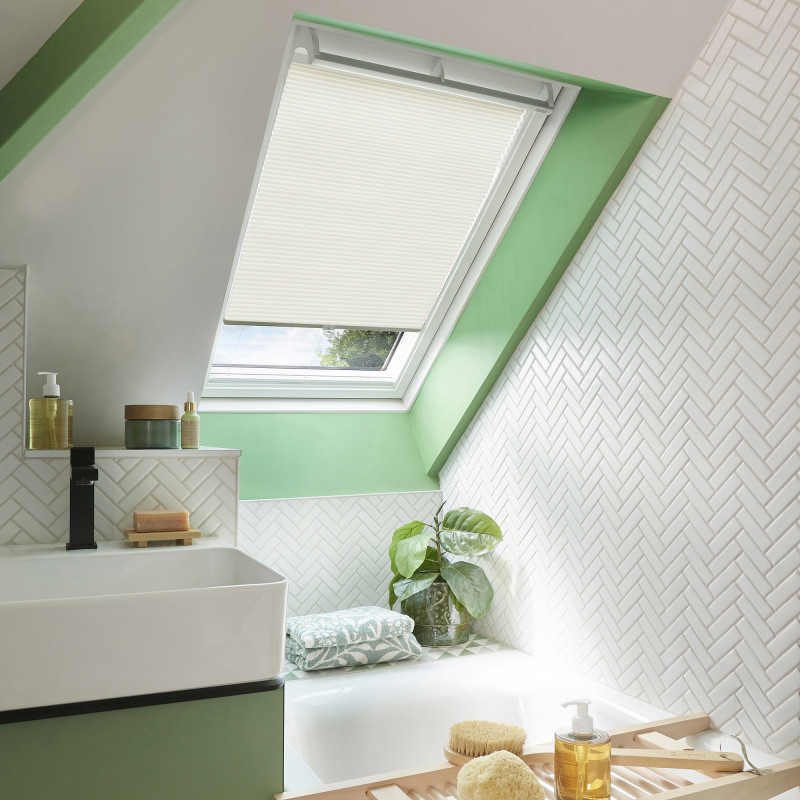 A white pleated skylight blind, mounted inside a roof window and pulled down partially, filtering daylight; bright sloped bathroom with green-painted trim, white herringbone tiles, sink, tub, plant.