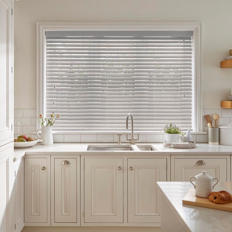 Horizontal white Venetian blinds lowered across a large window, slats mostly closed and gently filtering daylight; kitchen sink and marble countertop beneath, cream cabinets, small plants and utensils on counters.