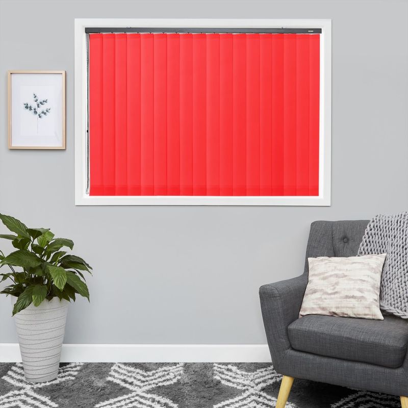 Red vertical blinds, mounted inside a white window frame with spaced slats, closed and blocking light; living room with grey walls, armchair, cushion and throw, potted plant, framed print.