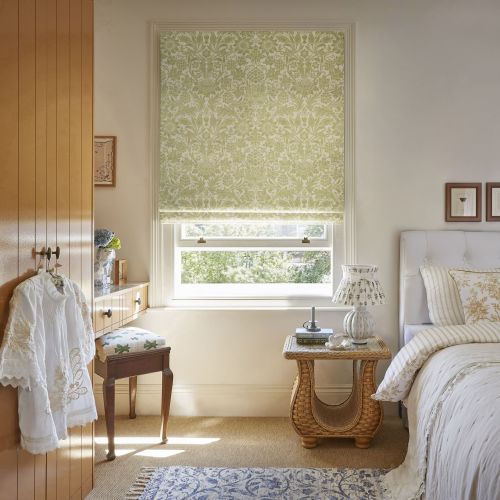 Floral-patterned Roman blind, mostly lowered, filtering soft daylight through a sash window; cozy sunlit bedroom with white bed, wicker bedside table, dressing table and leafy outside view.