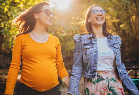 Two smiling women in sunglasses standing outside with the sun shining on them, one of the women is pregnant.