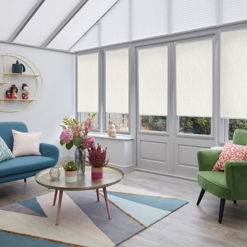 Cream-textured roller blinds across tall doors and windows, partially lowered and diffusing daylight; slanted roof pleated blinds closed; bright sunroom with teal sofa, green armchair, coffee table and flowers.