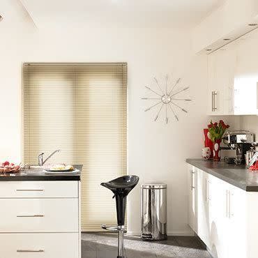 Beige horizontal Venetian blinds covering a central window, closed and softly filtering daylight; modern white kitchen with dark countertops, a black barstool, stainless bin, wall clock and tulip bouquet.