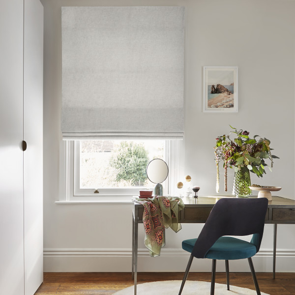 Pale grey Roman blind, fully lowered with gentle horizontal folds, filtering soft daylight into a home office. Wooden desk beneath holds mirror, vase of flowers, scarf; teal velvet chair.