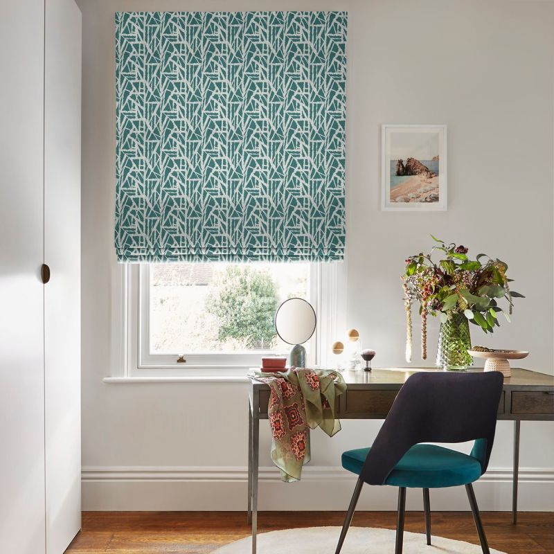 A teal Roman blind with white geometric pattern, lowered and filtering daylight through a sash window above a desk with chair, vase of foliage, mirror and print in a bright office.