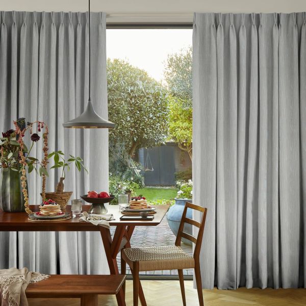 Floor-length grey pleated curtains, drawn slightly apart and gently filtering light; framing a sunlit dining room with wooden table set for breakfast and a view of a lush backyard garden.