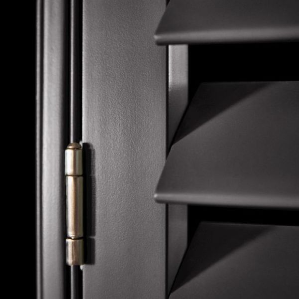 Matte dark louvered window shutters with angled horizontal slats and a brass hinge at left — slats closed, casting sharp shadows in a close-up interior view with low directional lighting.