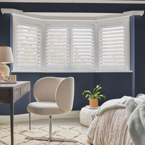 White plantation shutters fitted across a three-panel bay window, slatted panels tilted to filter daylight; set in a navy-walled bedroom nook with a cream chair, desk and potted plant.