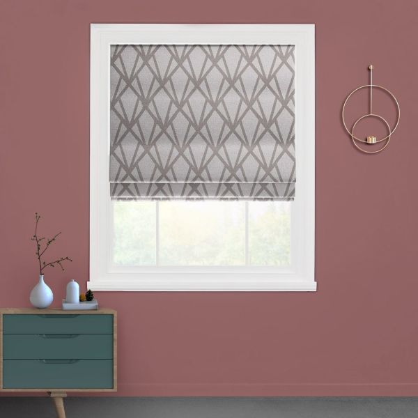 A grey geometric-patterned Roman shade lowered over a white-framed window, diffusing daylight; set in a mauve-walled room with a teal drawer, small vases on a console, and circular gold wall decor.
