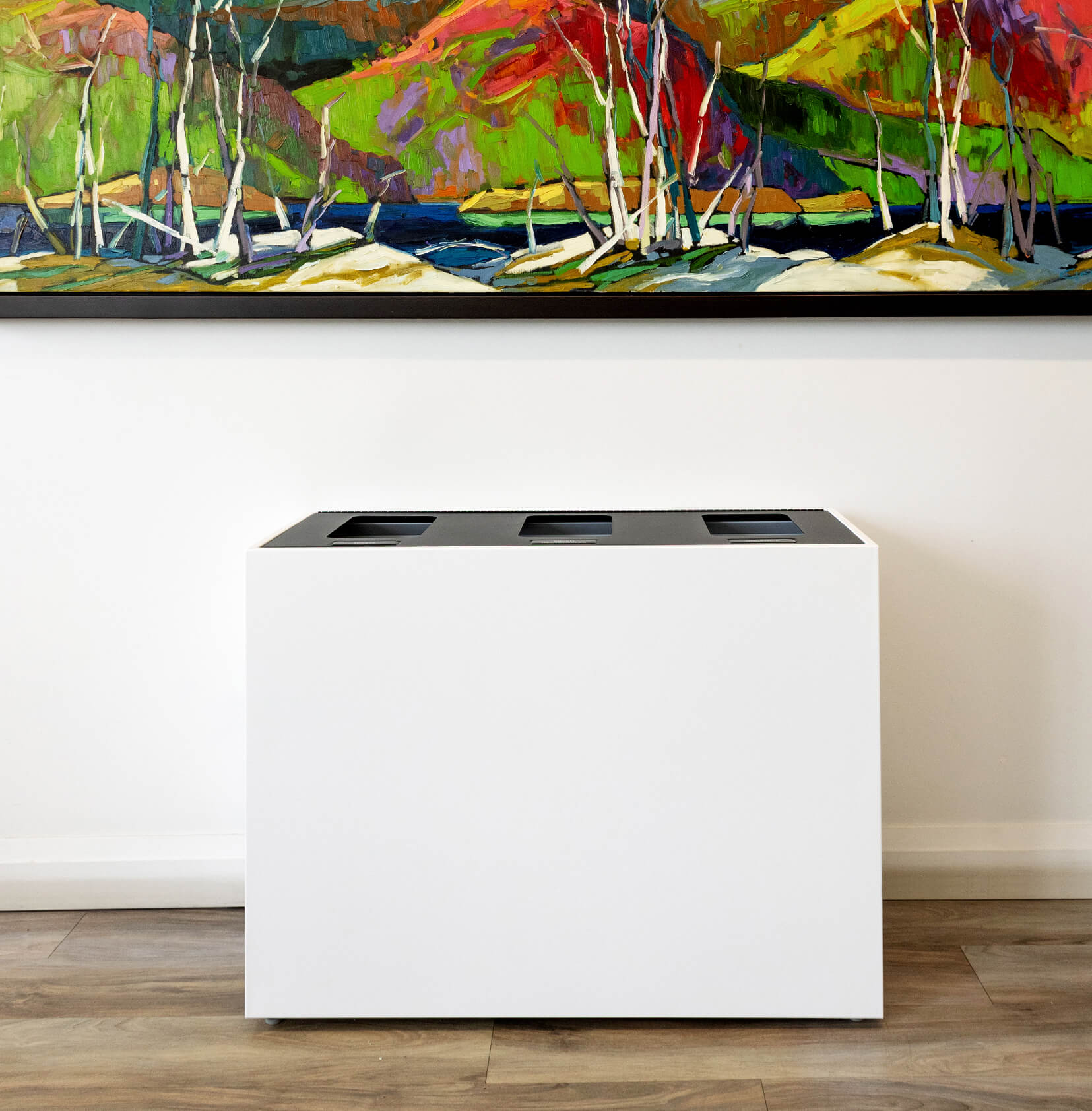 triple waste and recycling bin with a white body and black hinged lid sitting under a modern painting in an office hallway