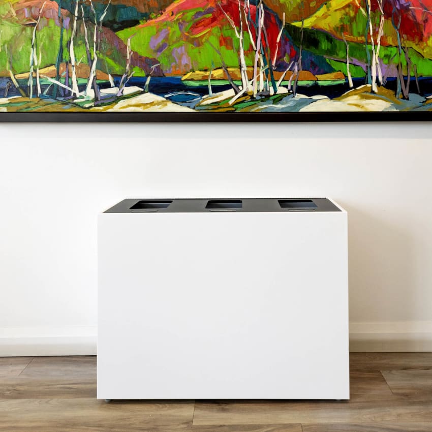 triple waste and recycling bin with a white body and black hinged lid sitting under a modern painting in an office hallway