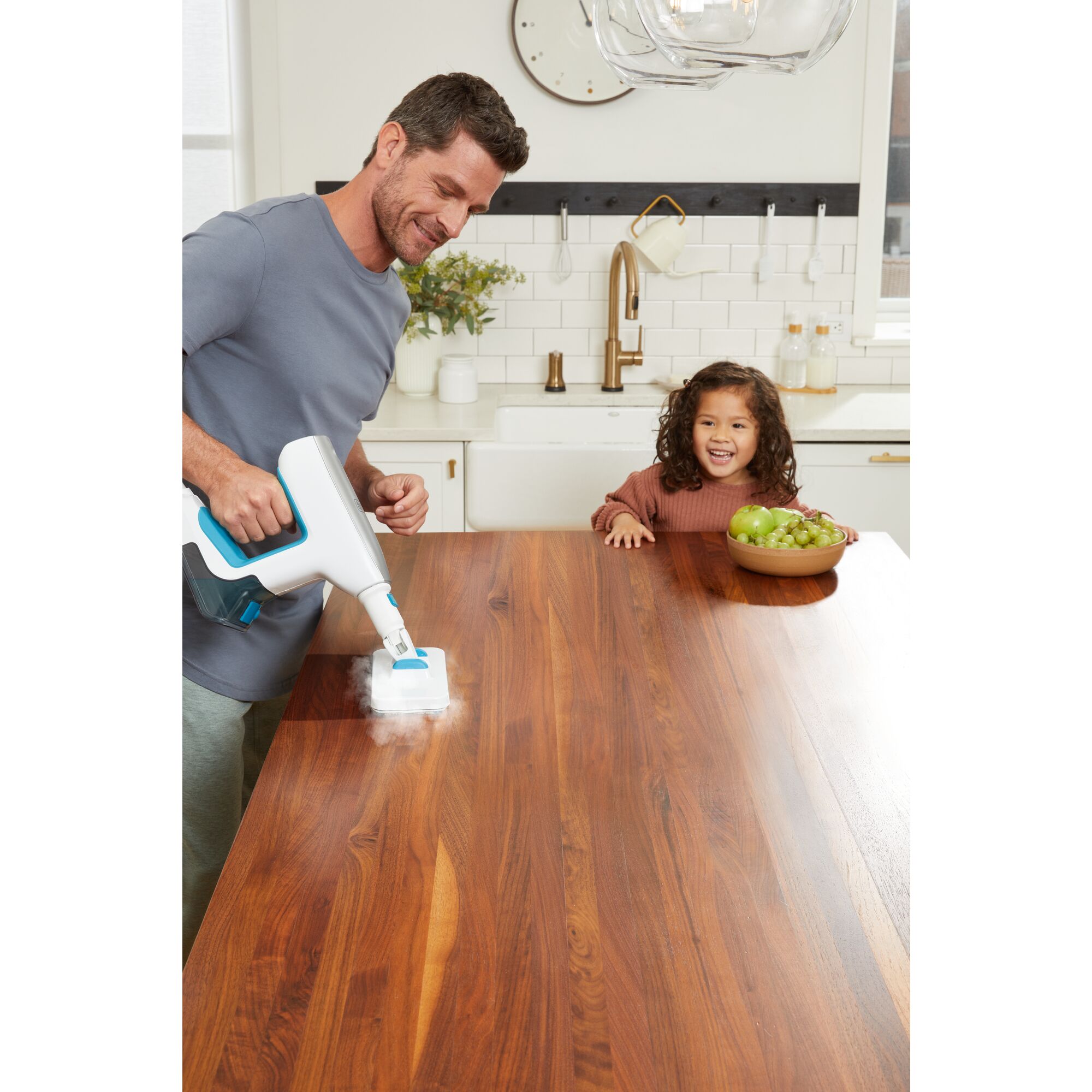 Man cleaning kitchen table with steam mop accessory while young child looks on