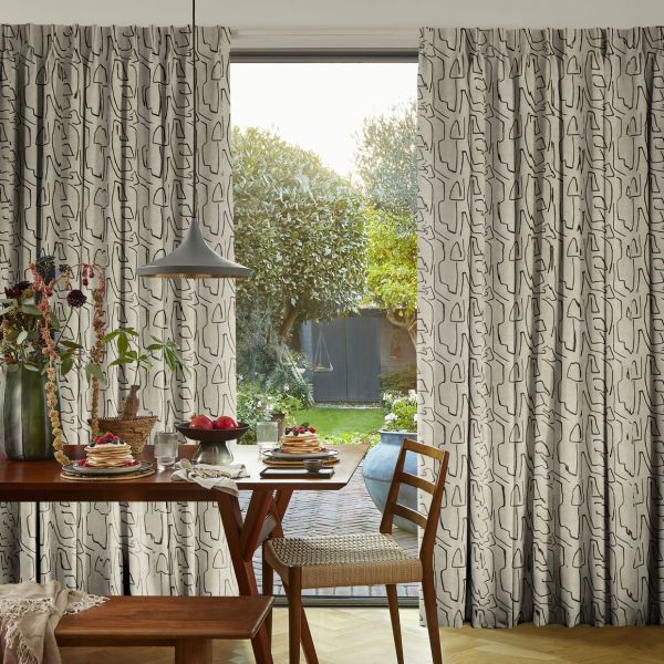 Pleated beige curtains with black abstract line pattern, drawn to sides and partially open, framing a sunlit backyard; wooden dining table set with pancakes, plates and pendant lamp in foreground.