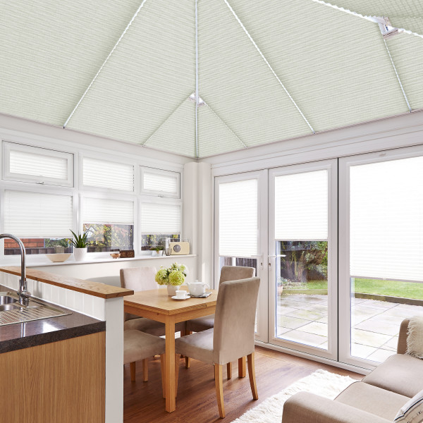 White pleated cellular blinds cover the pitched conservatory ceiling and all windows and doors, closed and softly diffusing daylight, in a bright kitchen-dining area with wooden table and garden view.