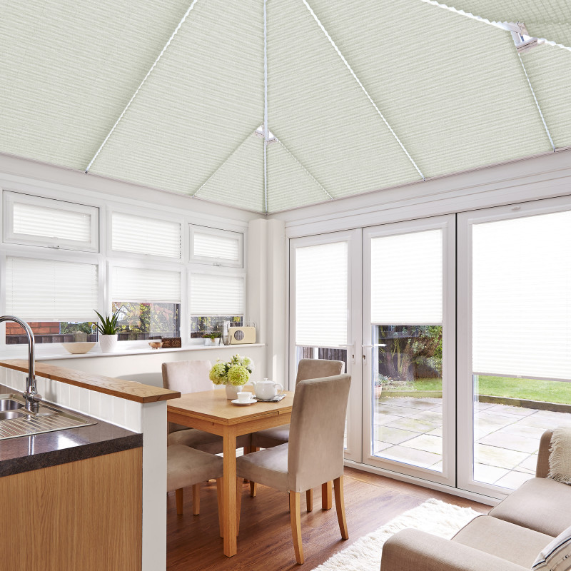 White pleated cellular blinds cover the pitched conservatory ceiling and all windows and doors, closed and softly diffusing daylight, in a bright kitchen-dining area with wooden table and garden view.