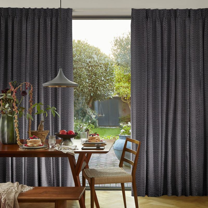 Charcoal-gray pleated curtains with textured weave, drawn partly open framing a glass door; filtering soft daylight into a dining room with a wooden table and leafy garden view.
