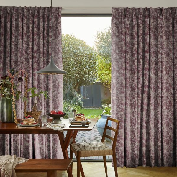 Full-length pleated plum-gray curtains, drawn aside to frame an open door and admit daylight; dining area with wooden table, pendant lamp, chair and leafy garden view.