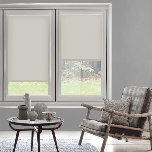 Beige fabric roller blinds covering twin windows â€” left fully lowered, right half-raised â€” filtering soft daylight into a neutral living room with a bamboo-frame armchair, coffee table, and garden view.