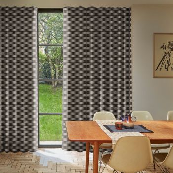 Floor-length patterned curtains with geometric print, drawn mostly closed leaving a narrow central gap showing a four-pane window; they filter soft daylight into a dining room facing a green garden.