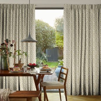 Beige diamond-patterned pleated curtains, full-length and parted at a sliding glass door, filtering soft daylight; wooden dining table set for breakfast in foreground, sunlit garden with trees and shed beyond.